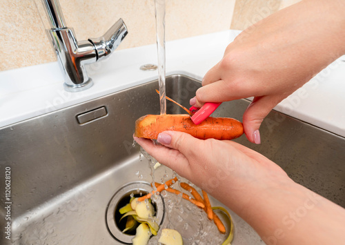 Peeling a fresh carrot under running water with waste collected in a garbage disposer.
