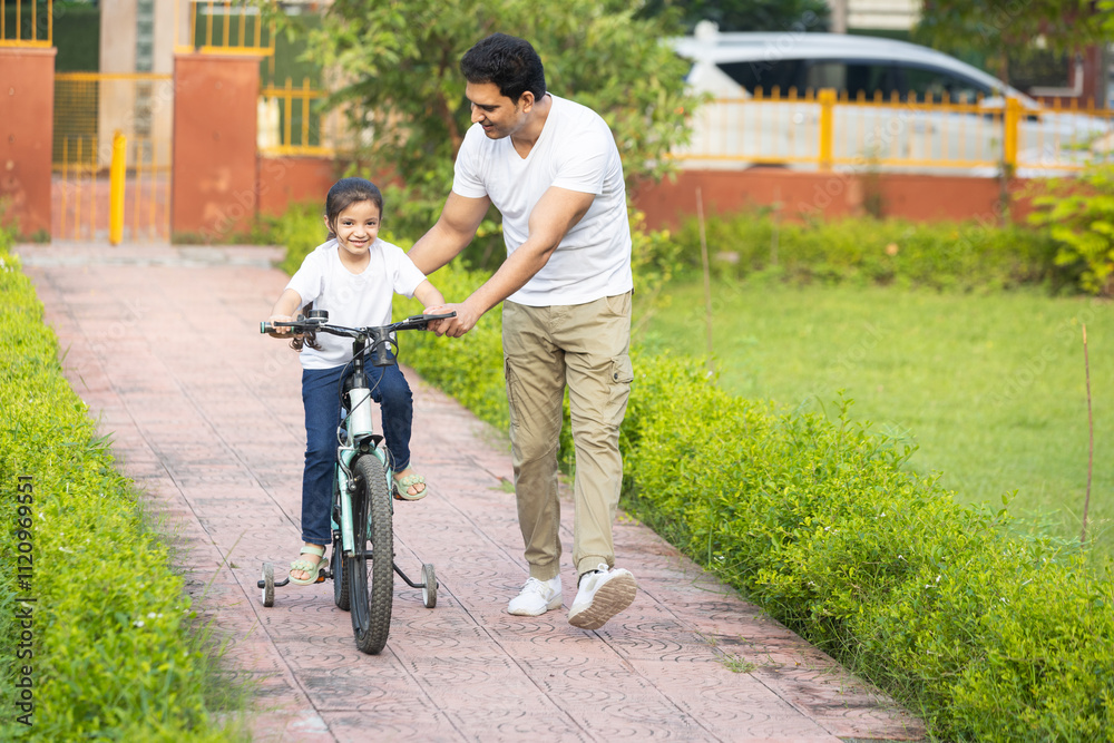 Young indian father teaching her daughter to ride bicycle in summer park outdoors. Dad helping and supporting her to cycle and pedal a bike. Young indian father teaching her daughter to ride bicycle in summer park outdoors. Dad helping and supporting her to cycle and pedal a bike.
