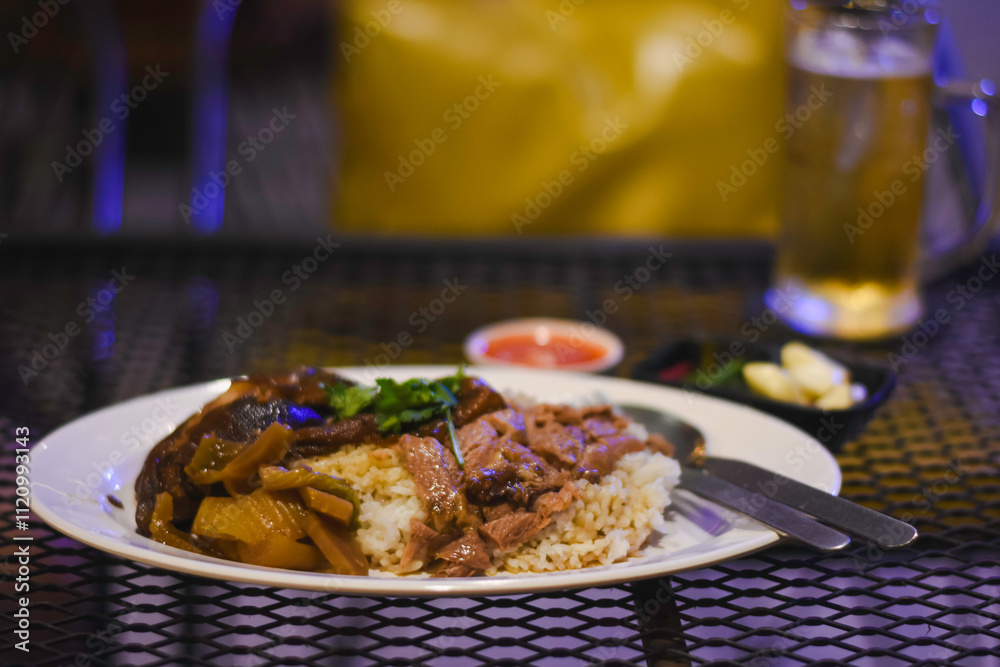 plate of food on a metal mesh table. The plate contains rice topped with what appears to be braised meat and vegetables, garnished with some herbs. plate of food on a metal mesh table. The plate contains rice topped with what appears to be braised meat and vegetables, garnished with some herbs.