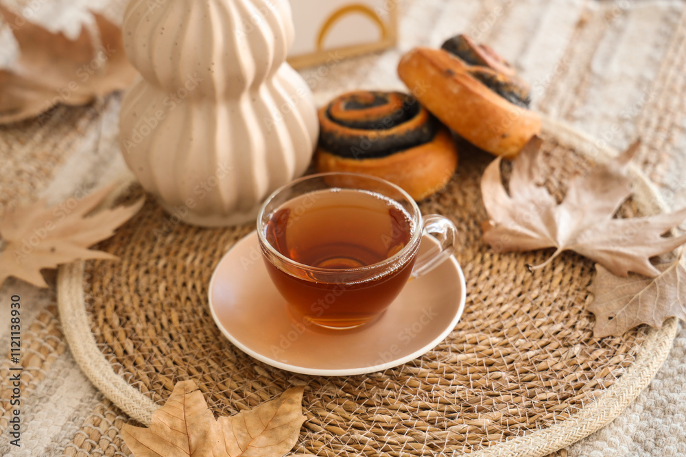 Wicker mat with cup of hot tea, tasty poppy buns and autumn leaves on table at home, closeup Wicker mat with cup of hot tea, tasty poppy buns and autumn leaves on table at home, closeup