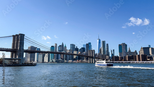 The iconic Brooklyn Bridge and New York City skyline on a sunny day with a ferry crossing the East River, symbolizing urban beauty.
