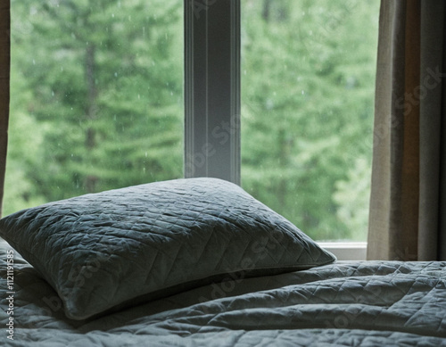 Photography A cozy bedroom scene with a soft pillow on a quilted bed near a window overlooking lush green trees
