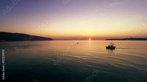 A serene sunset over calm waters with a boat in the foreground and hills in the background.