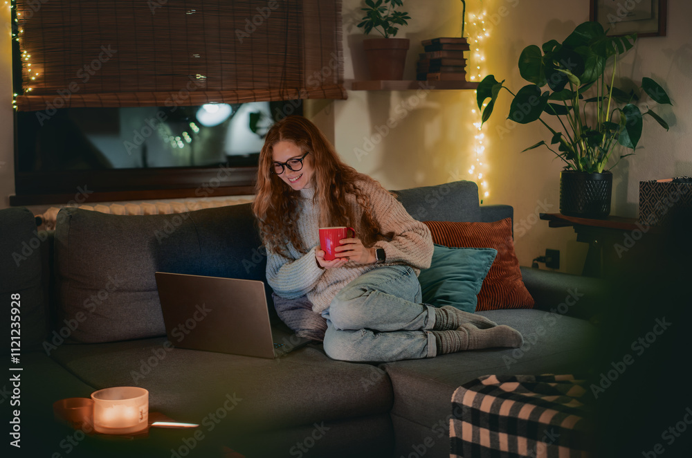 Obraz premium A young woman sitting comfortably on a couch using a laptop during a cozy and festive evening at home