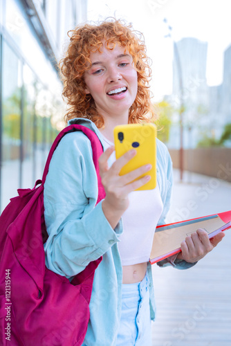 A young woman smiles while holding a smartphone and notebooks outdoors. She is dressed casually with a backpack, enjoying her time in a bright, modern environment with a positive, confident expression