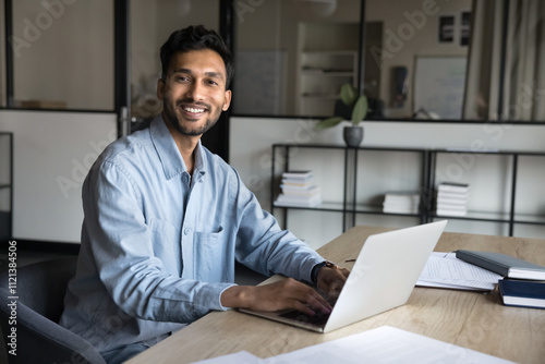 Businessman sits at workplace desk with laptop, smile, looks at camera, feels satisfied with career in company, professional achievements and opportunities. Workflow using tech, remote communication