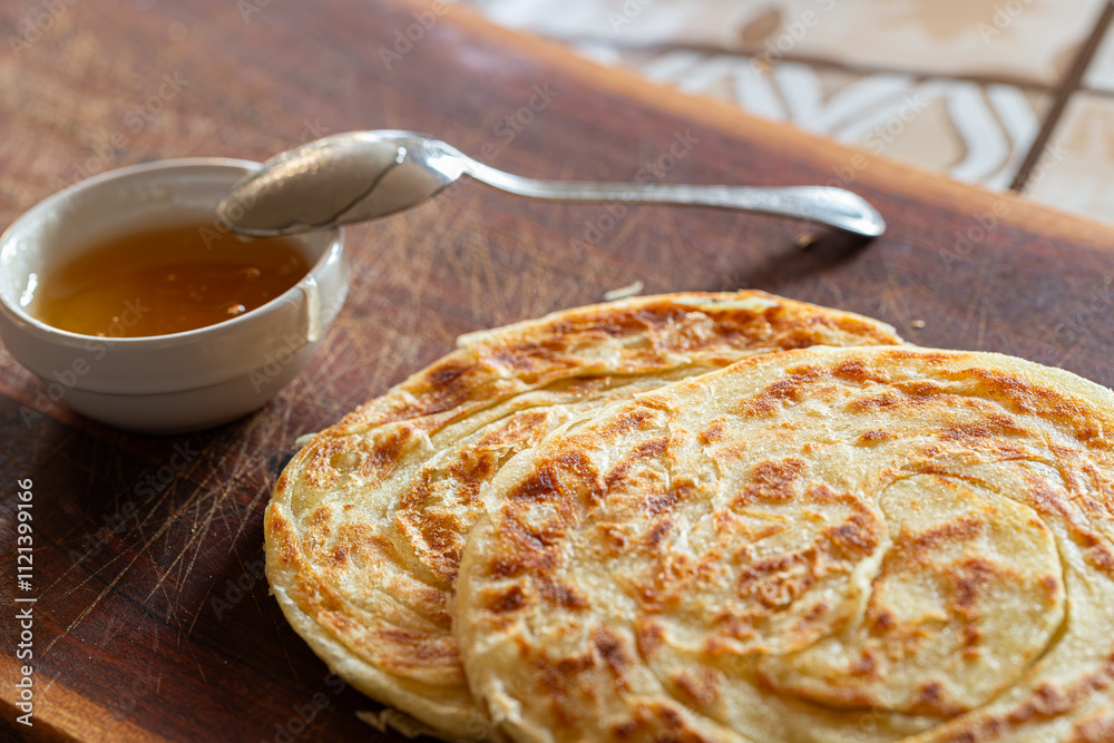 A plate of msemen on a wooden board, accompanied by honey in a white ceramic bowl and a silver spoon, offering a sweet Moroccan delight.