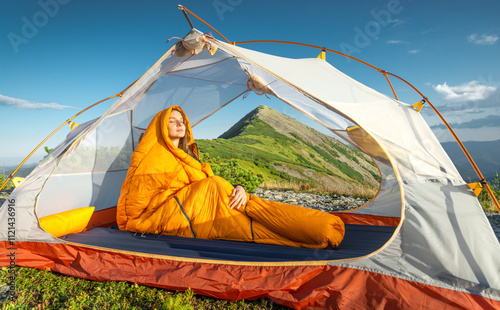 A young woman in a yellow sleeping bag with her eyes closed, enjoying the warmth of the sunset from inside her tent. photo