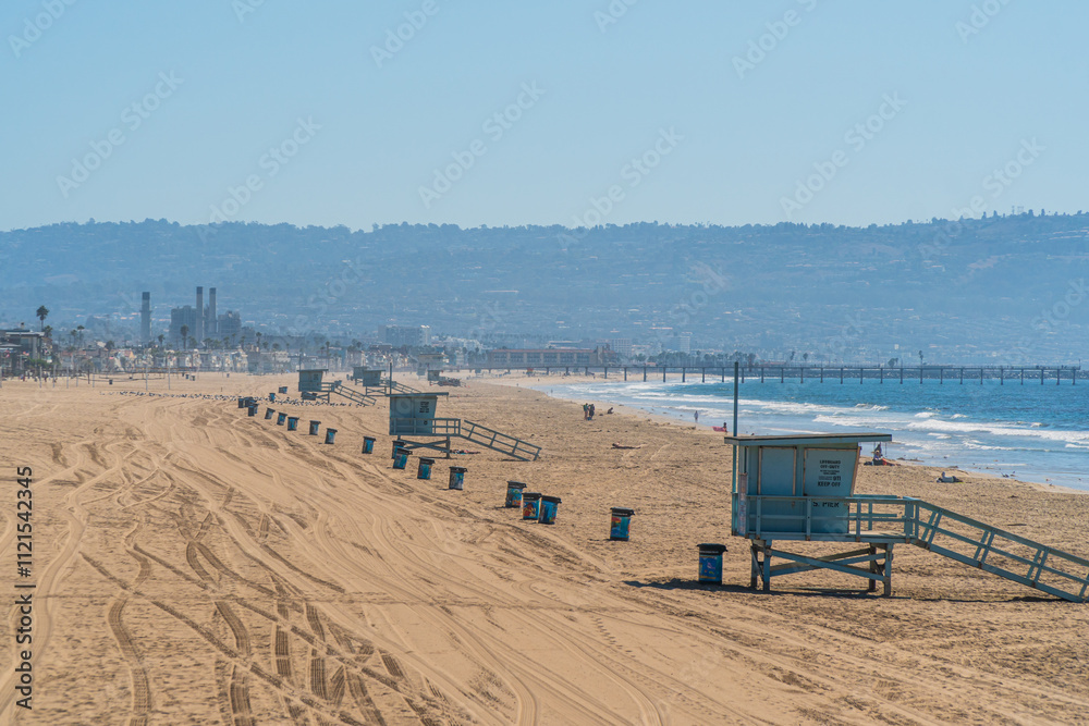 Obraz premium Californian beach near Los Angeles with typical lifeguard huts and garbage bins aligned on the sand, and El Segundo power plant in the distance