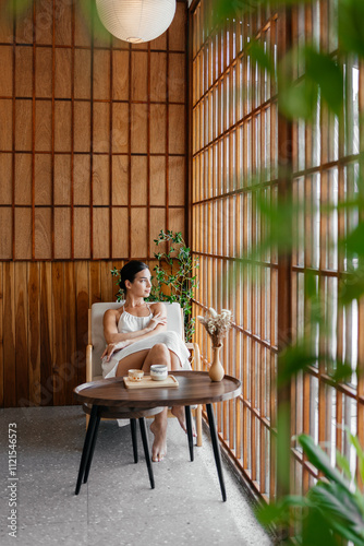 Young, caucasian woman enjoying a cup of coffee, tea lounge chilling near window in modern cozy apartment. Relaxed serene brunette girl feel fatigue lounge on comfortable sofa at home.