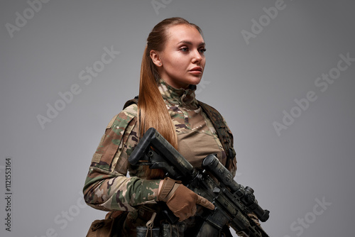 A confident woman in tactical military gear holds a rifle, exuding strength and readiness. Dressed in camouflage with a tactical vest, gloves, and protective headphones, she embodies focus and profess