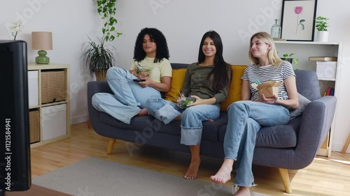 Three happy young beautiful female friends watching TV while eating heathy food sitting on the sofa at home. Leisure, friendship and domestic lifestyle concept. photo