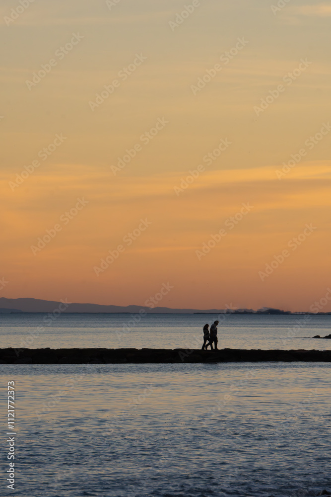 Silhouette of a couple walking by the Mediterranean sea on a pier breaking waves in a reddish summer sunset orange clouds and silhouette of mountains in the background. Valencia Spain. Silhouette of a couple walking by the Mediterranean sea on a pier breaking waves in a reddish summer sunset orange clouds and silhouette of mountains in the background. Valencia Spain.