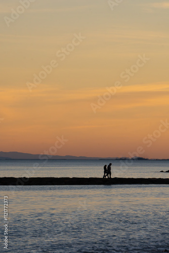 Silhouette of a couple walking by the Mediterranean sea on a pier breaking waves in a reddish summer sunset orange clouds and silhouette of mountains in the background. Valencia Spain.