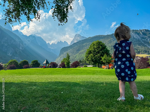 Fotografie Female toddler in beautiful dress walking on lush green meadow in Molveno with scenic view of mountains of majestic Brenta Dolomites, National Park Adamello Brenta, Trentino, Italy