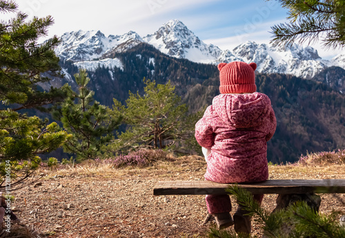 Small child sitting on wooden bench on top of Monte Nebria Est with panoramic view of snow capped mountains of Julian Alps, Friuli Venezia Giulia, Italy. Family hike in alpine wilderness in spring photo