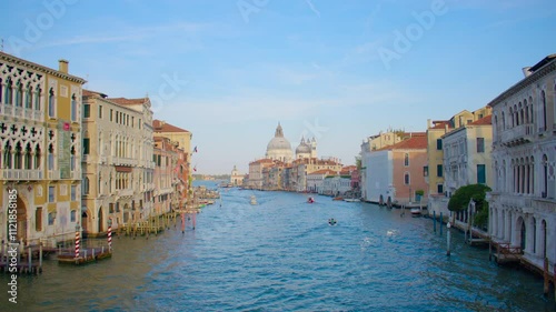 A vibrant view of Venice's Grand Canal, with ornate buildings on both sides. In the distance, the domed Basilica di Santa Maria della Salute stands under a clear blue sky, with boats dotting the canal