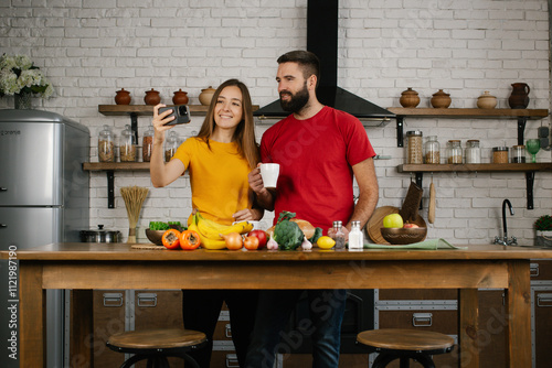 A young attractive man and a woman are standing in a kitchen and looking at cell phone in front of them. Lovely couple preaparing breakfast and looking in mobile phone in the kitchen at home. photo