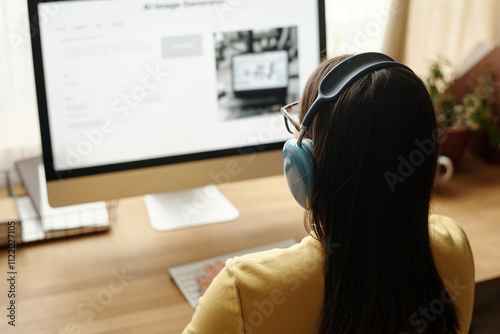 Focus on rear view of young woman with dark long hair listening to music in headphones in front of computer photo