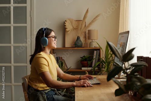 Young smiling woman in casual apparel and headphones sitting by workplace in front of desktop computer