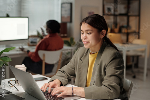 Young smiling businesswoman in formalwear looking at laptop screen and typing on keypad in coworking space photo