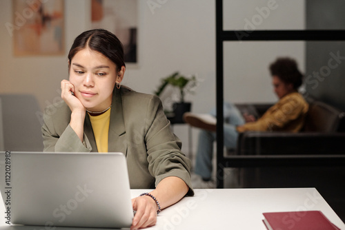Young smiling female office worker looking at laptop screen while watching online video in coworking space