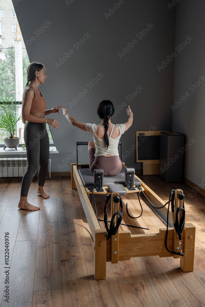 Fototapeta premium Woman training pilates on the reformer bed with trainer. Reformer pilates studio machine for fitness workouts in gym. Fit, healthy and strong authentical body. Fitness concept