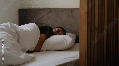 A young handsome Caucasian bearded man is sleeping sweetly on a bed at home under a white blanket. The guy is resting in his apartment lying on the bed alone, wakes up and yawns