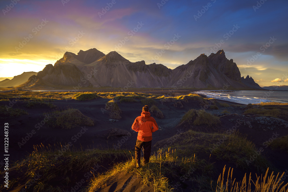 Fototapeta premium A hiker in a red rain jacket stands on a grassy sand dune at sunset, admiring Vestrahorn and Klifatindur in Stokksnes, Iceland.