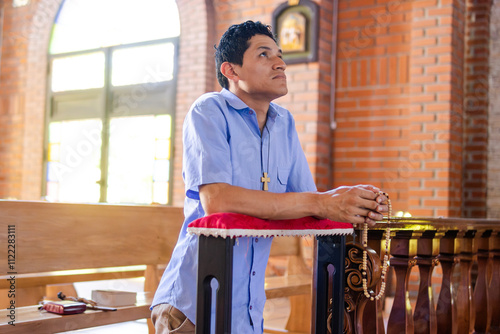 A young Latino man kneeling with a rosary in his hand, looking upward, radiating peace and tranquility in his prayer. Horizontal composition with copy space