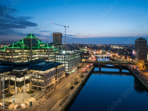 A dusk view of Dublin's modern skyline with the iconic green-lit Bank of Ireland building, a river reflecting the city lights, and a bridge crossing over it.