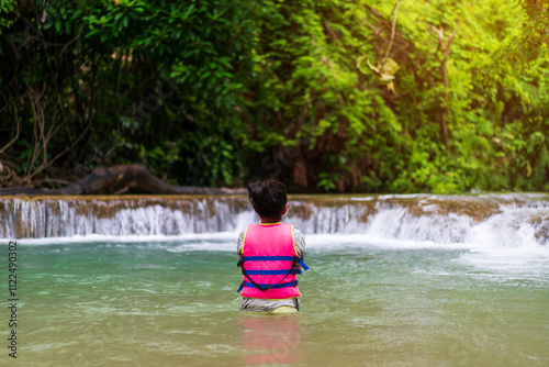 back asian child standing in waterfall or kid girl wearing life jacket to people playing stream water on nature in forest wild garden with trees on summer holiday travel at huai mae khamin waterfall