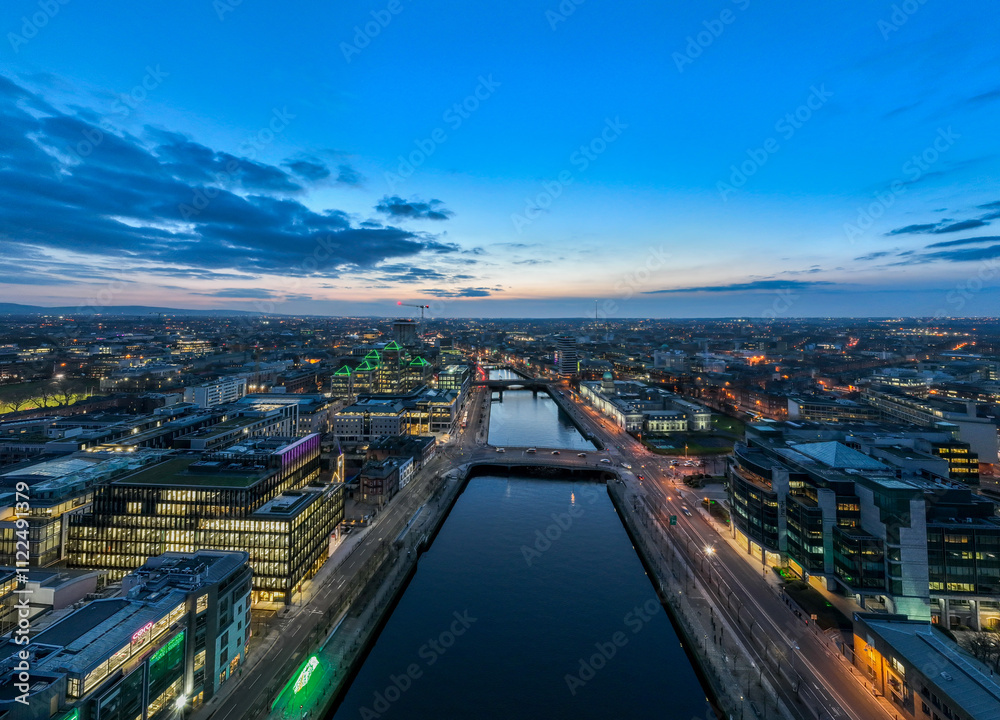 Fototapeta premium A dusk view of Dublin's modern skyline with the iconic green-lit Bank of Ireland building, a river reflecting the city lights, and a bridge crossing over it.