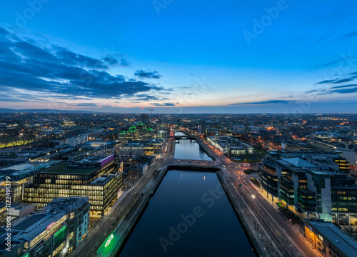 A dusk view of Dublin's modern skyline with the iconic green-lit Bank of Ireland building, a river reflecting the city lights, and a bridge crossing over it. photo