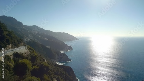 Scenic coastal highway winding through a mountainous terrain, overlooking a tranquil ocean. Sunlight reflects on the water's surface. photo