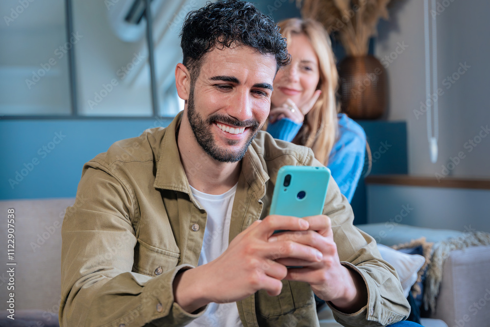 Fototapeta premium Smiling man using smartphone with a woman in the background at home