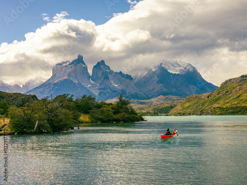 A vibrant red canoe glides across turquoise waters in Chilean Patagonia, surrounded by rugged mountains, lush greenery, and dramatic clouds in Torres del Paine.