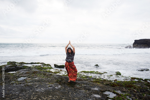 Asian woman wearing native Sarong of Bali doing yoga poses on the coast of the Indian Ocean at Bali Island of Indonesia. Travel destination and freedom life of Asian concept photo