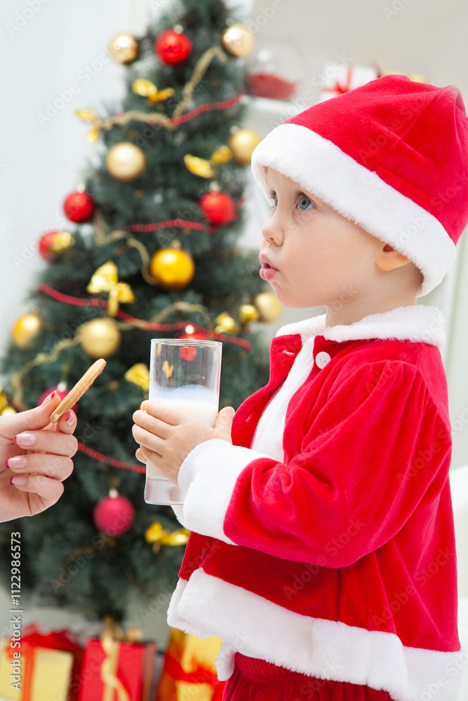 Portrait of little cute boy in Santa hat drinking milk with biscuit