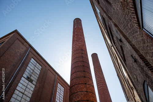 Low-angle view of tall brick factory chimneys and industrial buildings against a clear blue sky.  Brickwork and windows are prominent features. photo