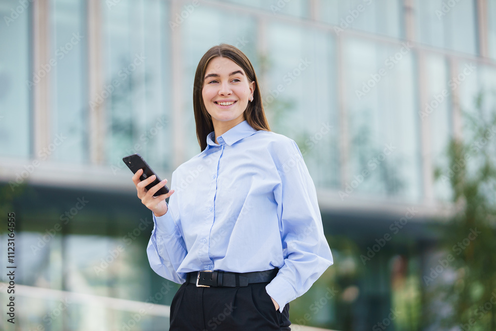 Fototapeta premium Young woman using smartphone while standing outside a modern building on a sunny day