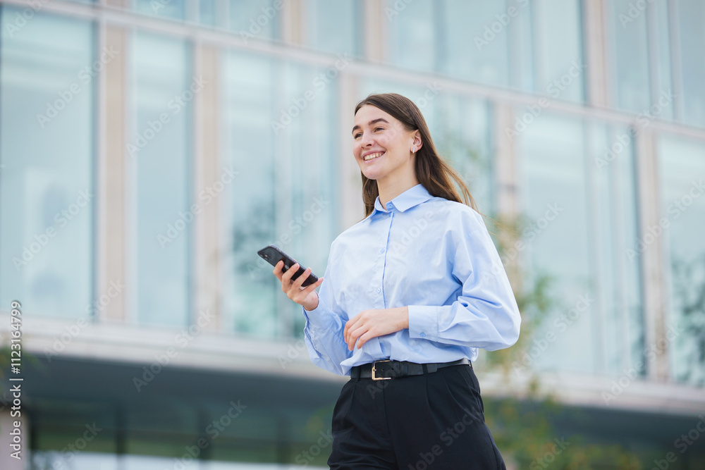 Young woman walking outside while using smartphone near modern building in bright daylight Young woman walking outside while using smartphone near modern building in bright daylight