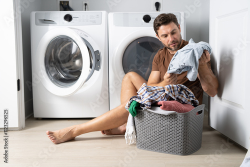 Happy man sorting clothes near washing machine in laundry room. man sits on the floor of a house near a washing machine with dirty clothes. a man does his laundry at home. a man holding dirty clothes