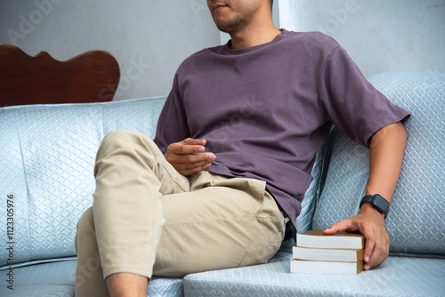 A man is seated comfortably on a sofa, engaged in reading a book. His attire is casual yet neat, such as a shirt and trousers, suggesting a moment of leisure within a serene environment.