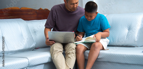 father and son are seated together on a comfortable sofa, collaborating on homework. The boy holds a book, while the father uses a laptop to complement their study session.education and family concept