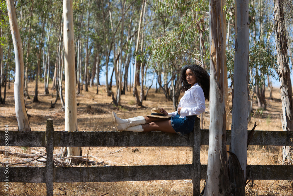 Fototapeta premium woman, young, beautiful, Latin, brunette with curly hair dressed in white shirt, denim skirt, white boots and hat, sitting with her legs stretched out on the forest fence. The woman rests.