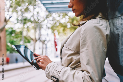 Cropped side view of young woman in trendy coat checking mail and reading notification on touch pad using free 4G internet.Pensive female blogger updating profile on tablet standing on tram stop photo