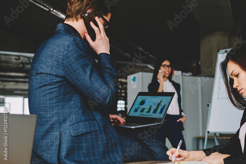 Caucasian female writing organisation plan in textbook while male economist analyzing graphic on laptop device and making important telephone conversation, concept of collaboration and communication