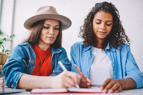 Clever hipster girls doing school homework together writing answers in textbook for education, young multicultural female persons notes organisation plan of course work sitting in cafeteria photo