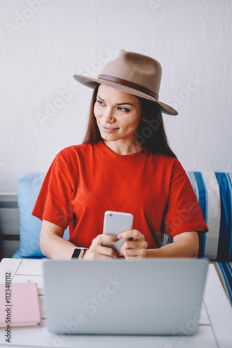 Positive female student in trendy hat enjoying time for e learning via laptop computer in cafeteria using free wifi connection, successful hipster girl holding modern smartphone gadget in hands photo